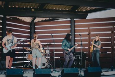 A band performs live on an outdoor stage with guitars, drums, and a saxophone.