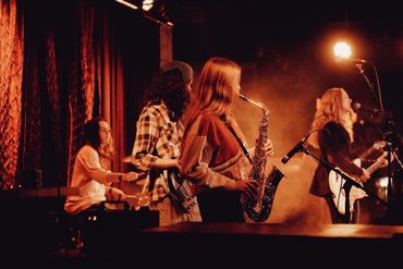 A live band performs on stage with saxophone, guitar, and drums under warm lighting.