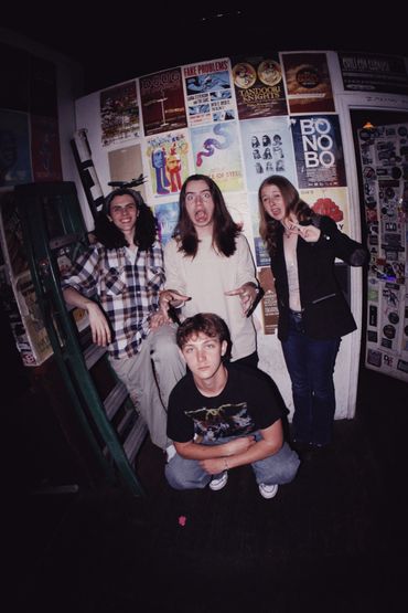 A group of four young people posing playfully in a room with poster-covered walls.