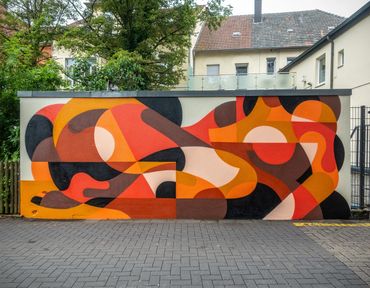 Colorful abstract mural with overlapping shapes in orange, red, black, and brown on an outdoor wall.