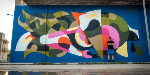 A man stands in front of a large colorful abstract mural on a blue wall.