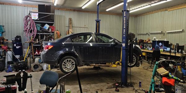 A black car is elevated on a lift inside a cluttered auto repair shop.