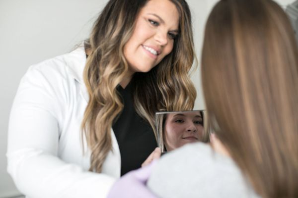 A smiling woman in a white coat interacts with another woman holding a mirror.