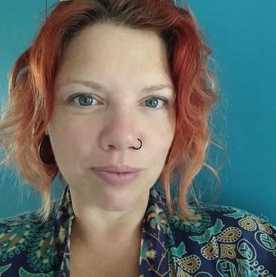 A picture of the website owner, she has red wavy hair, blue grey eyes, a nose piercing and full lips.