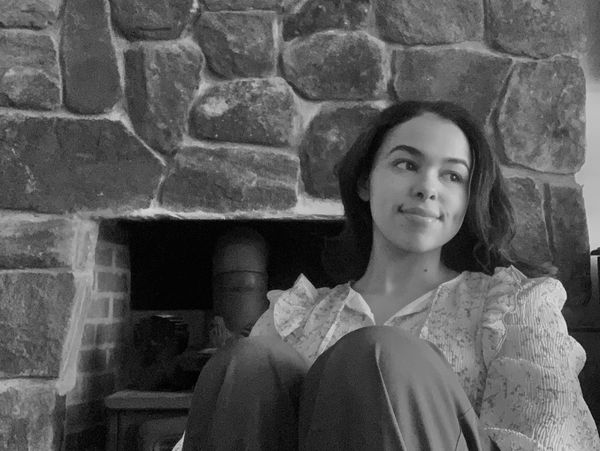Young woman sitting thoughtfully near a stone fireplace in black and white.
