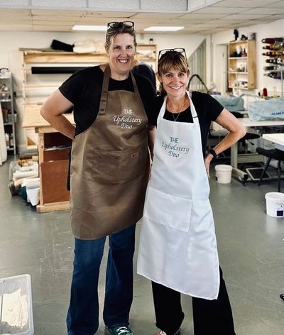Two women smiling in aprons labeled 'The Upholstery Duo' in a workshop.