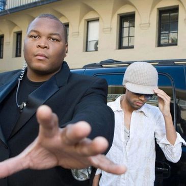 Two men posing outside, one reaching forward, the other adjusting a hat.
