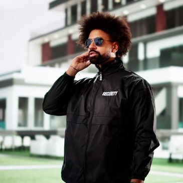 Man wearing a black security jacket and sunglasses outdoors.