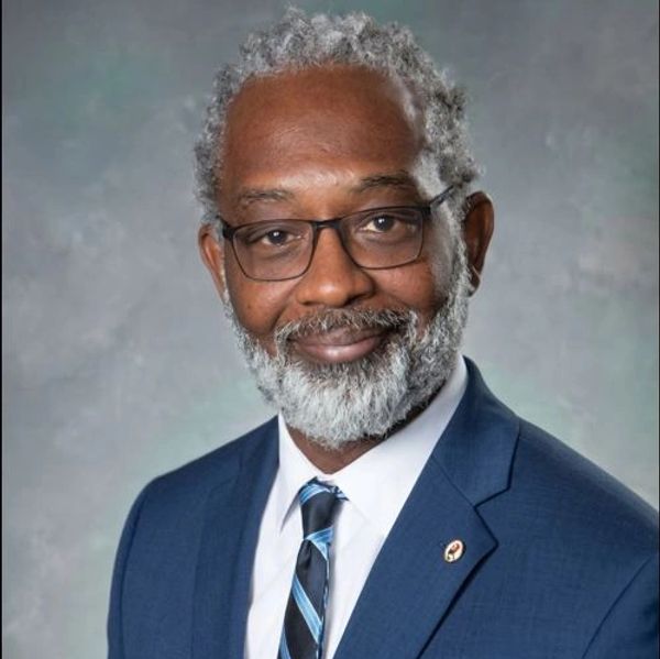 Smiling man with gray hair and beard in blue suit and glasses.