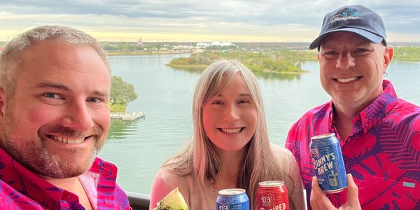 Three people smiling with tropical drinks and cans, overlooking a lake with a distant amusement park.