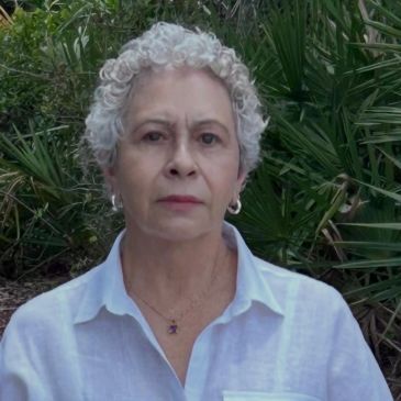 Mujer mayor con cabello rizado gris, vestida con camisa blanca, al aire libre.