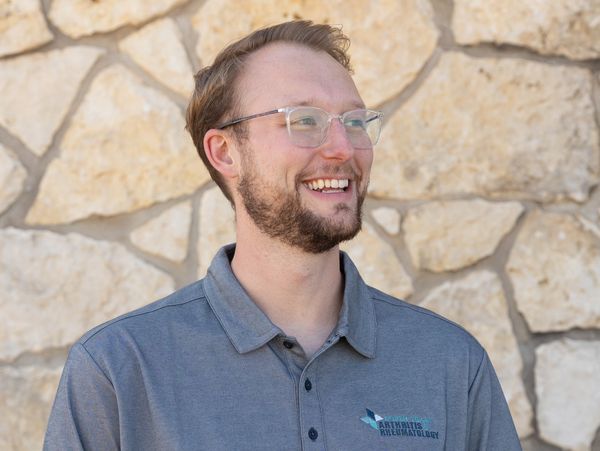Smiling man in glasses and gray polo with stone wall background.