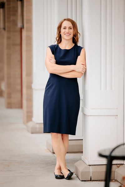 smiling woman in a blue dress leaning on a white pillar with arms crossed in front of her