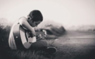 A young boy playing an acoustic guitar outdoors, sitting on the grass.