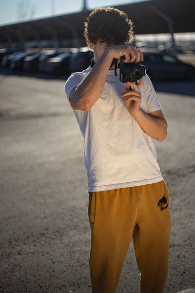 Young person in white shirt and yellow pants taking a photo outdoors.
