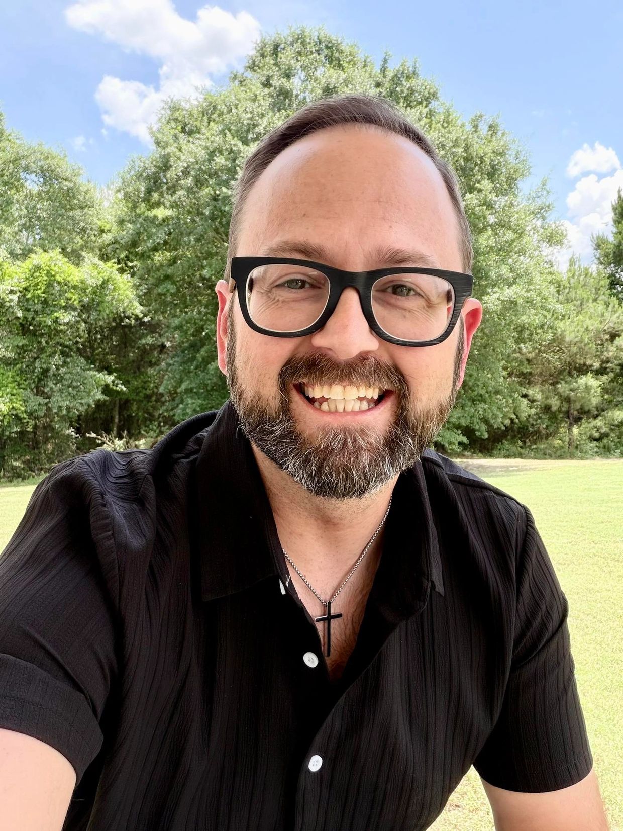 Smiling man with glasses and beard outdoors on a sunny day.