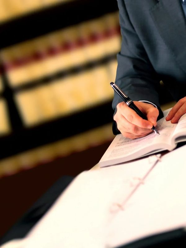Person in suit writing notes in a book with blurred library shelves in background.