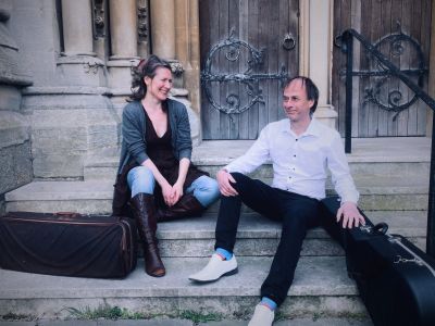 Two musicians sit on stone steps outside a historic building with their instrument cases.