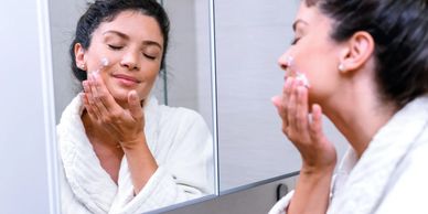 Woman in a white robe applying face cream in front of a bathroom mirror.