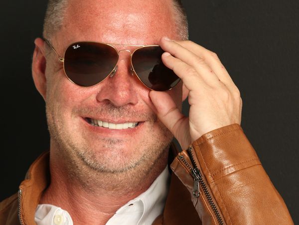 Man wearing brown leather jacket and Ray-Ban sunglasses, smiling.