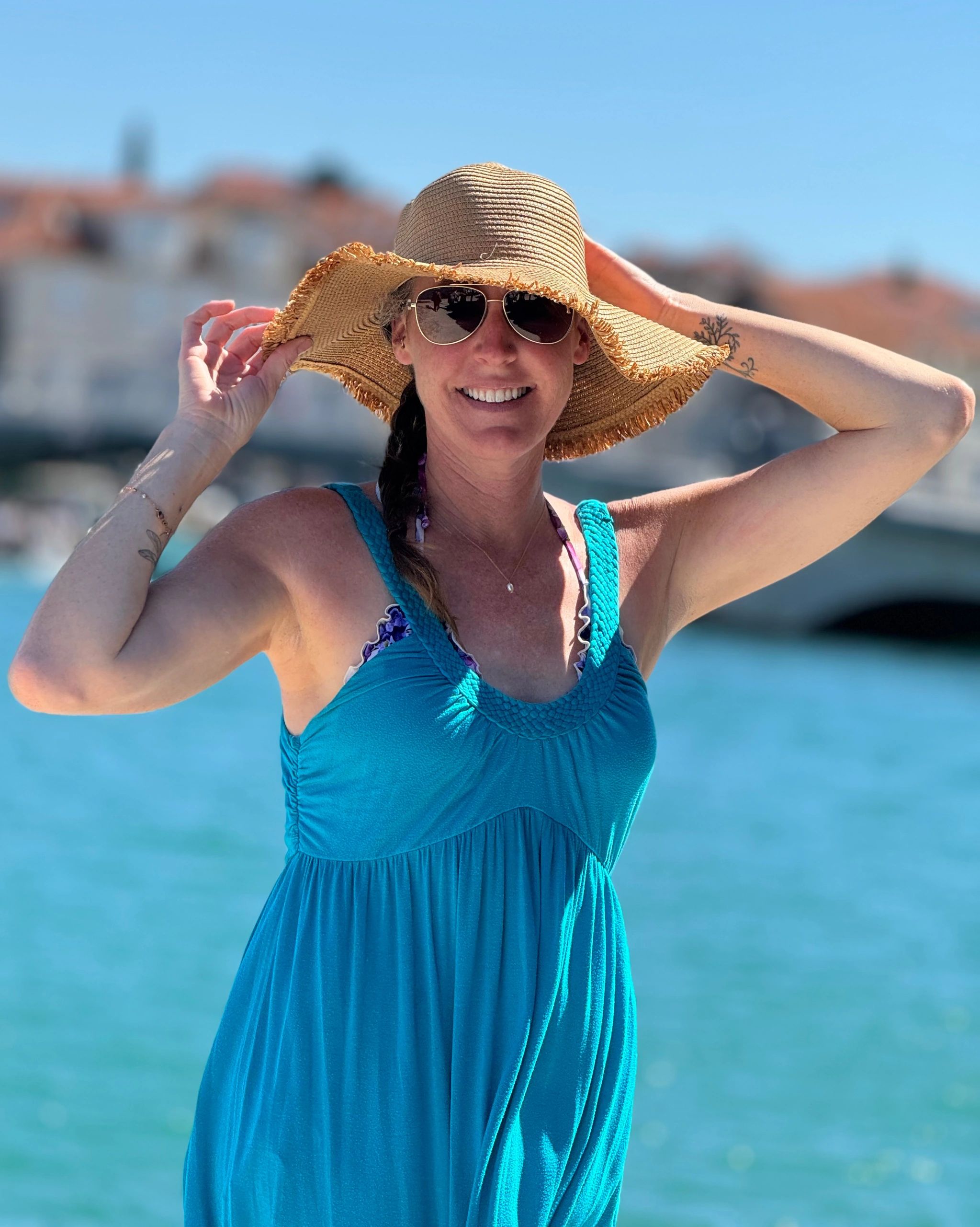 Smiling woman in a blue dress and sunhat by the water on a sunny day.