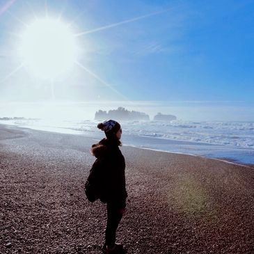 Person in winter clothing standing on a beach under the bright sun.