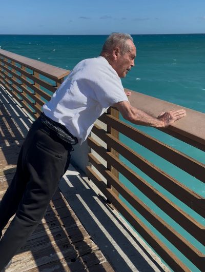 Elderly man leaning on a pier railing, looking at the ocean.