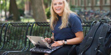 smiling blond female student sitting outside on a bench with laptop on her lap