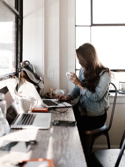 Woman with long brown hair sipping coffee and working at a laptop in a sunny coffee shop window