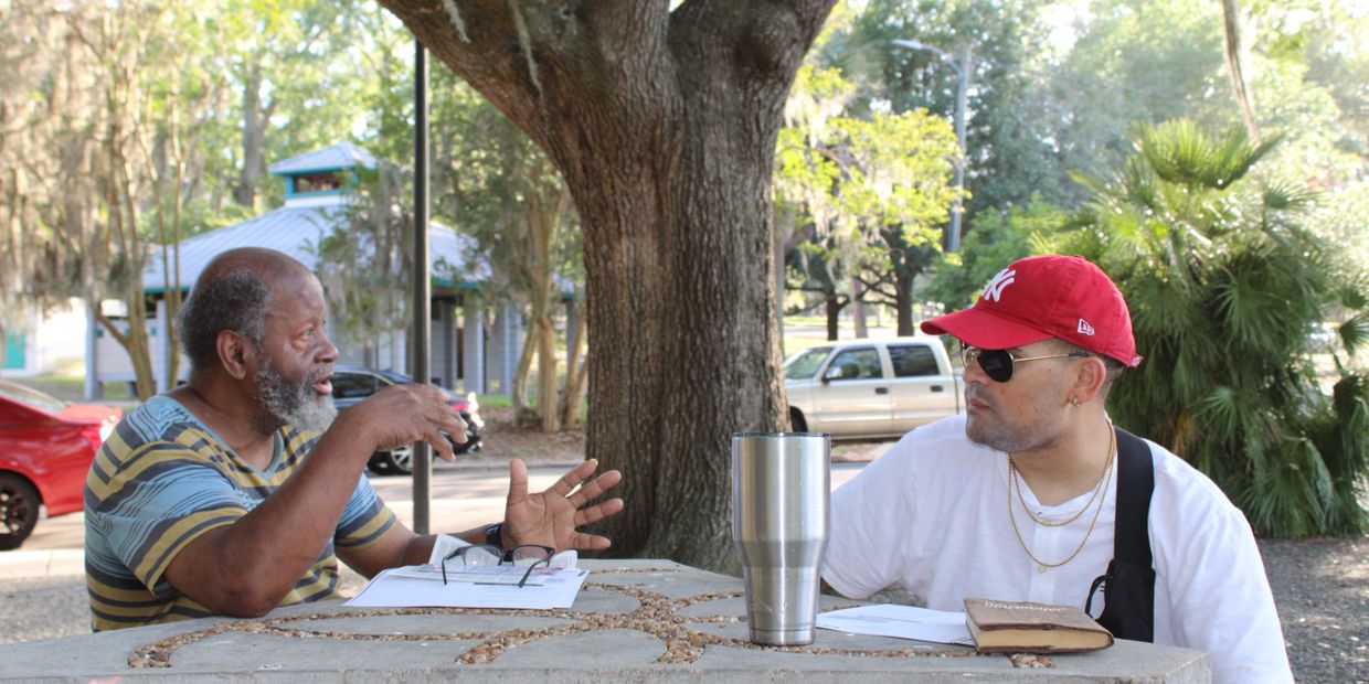 Two men having a conversation outdoors at a stone table under a tree.