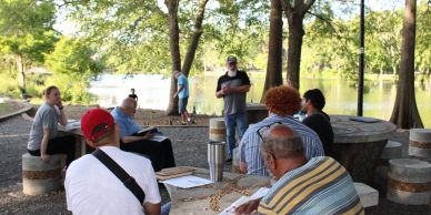 People gathered outdoors around a circular stone table near a lake, engaging in conversation.