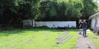 Two men standing by a house with ladders on the grass and a wooded area behind.