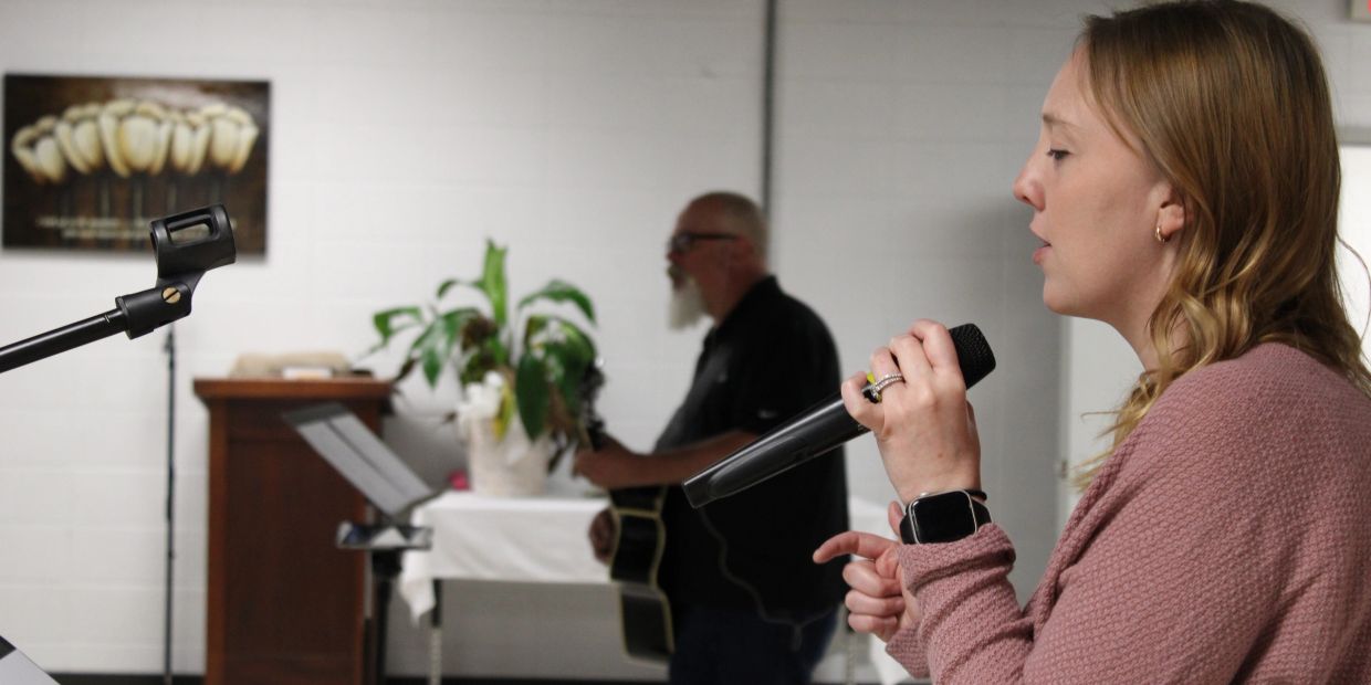 A woman singing into a microphone with a man playing guitar in the background.