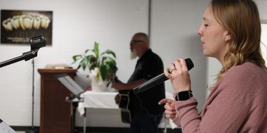 A woman sings into a microphone while a man plays guitar in the background.