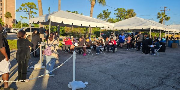 People wait in line and sit under tents in a sunny outdoor area.