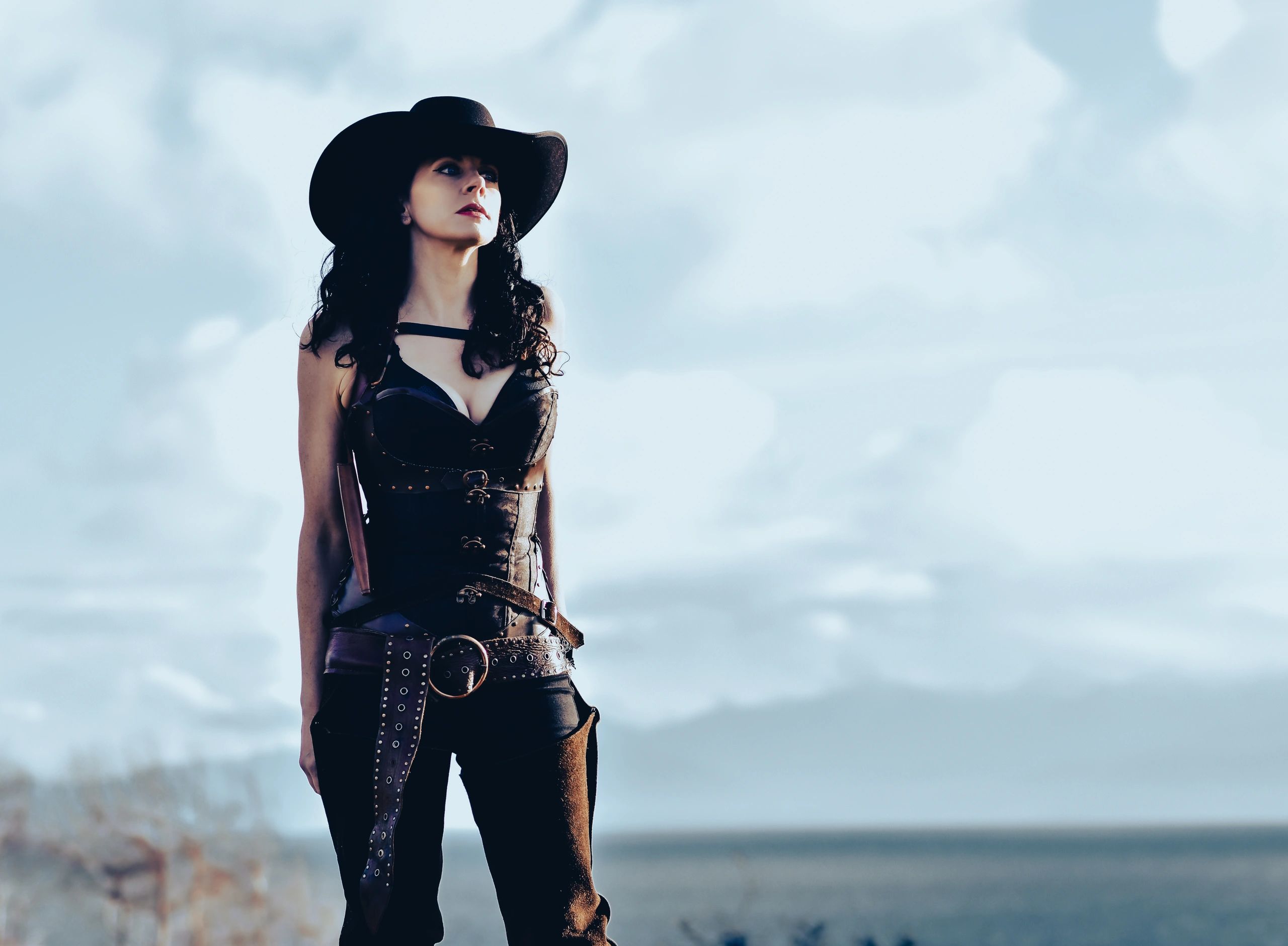 Woman in cowgirl get-up and cowboy hat standing in front of a blue sky backdrop 