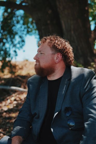 Man with curly hair and beard in blazer looking sideways outdoors.