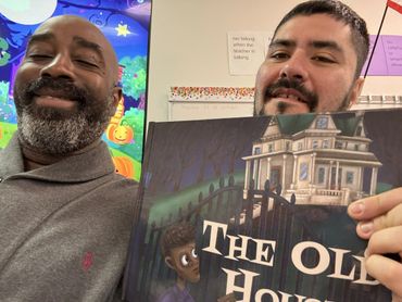 Two men smiling, holding a book titled 'The Old House' in a colorful classroom.