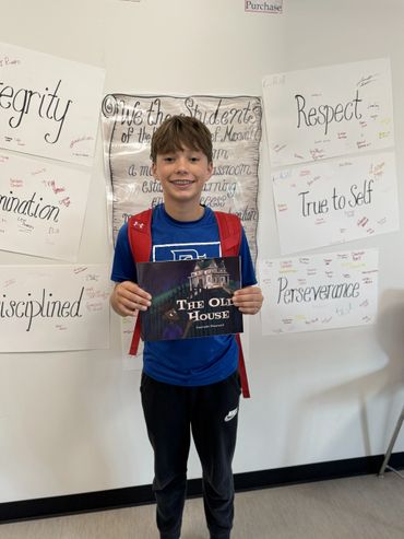 A smiling boy in a blue shirt holding a book titled 'The Old House' in a classroom.
