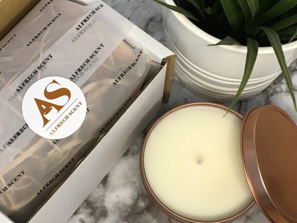 Alfrech Scent candle in a rose gold tin next to a packaged candle and decorative plant.