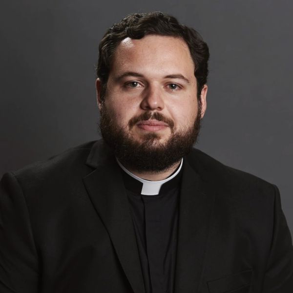 Portrait of a man in a clergy collar and black suit against a gray background.