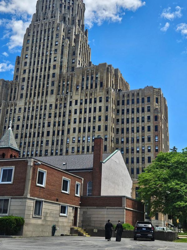 Two people walk near a tall art deco skyscraper under a blue sky.