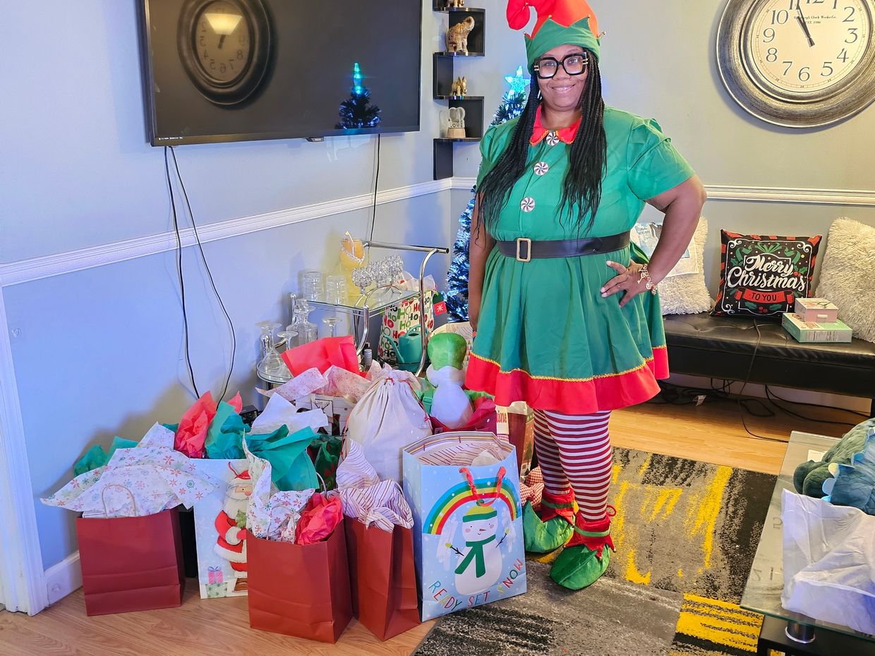 A woman dressed as a Christmas elf stands beside festive gift bags in a decorated living room.