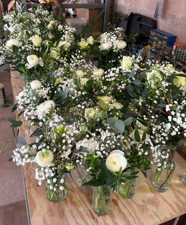 Multiple vases with white flowers and greenery arranged on a wooden table.