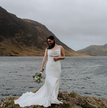Bride in a white dress stands by a lake with mountains in the background.