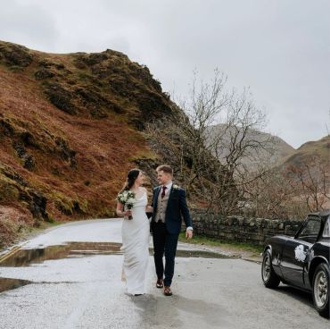 Newlywed couple walking happily on a mountain road beside a vintage car.