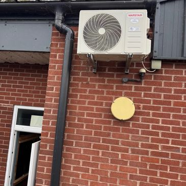 Outdoor air conditioning unit mounted on a brick wall beside a black drainpipe.