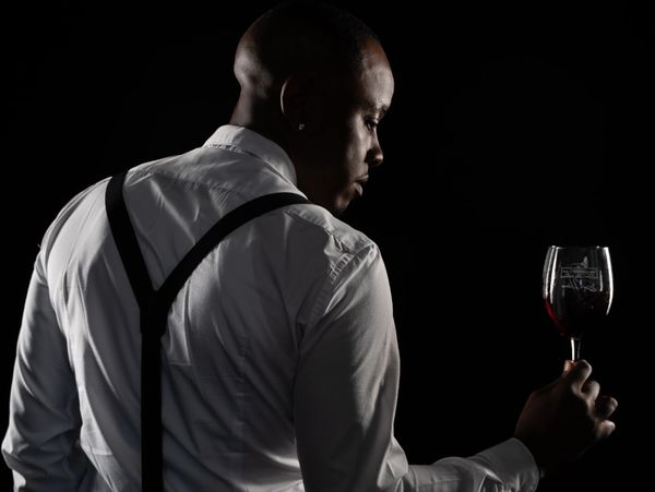 Man in white shirt with suspenders holding a wine glass against a black background.