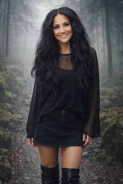 Smiling woman with long black hair in a black outfit stands on a forest path.