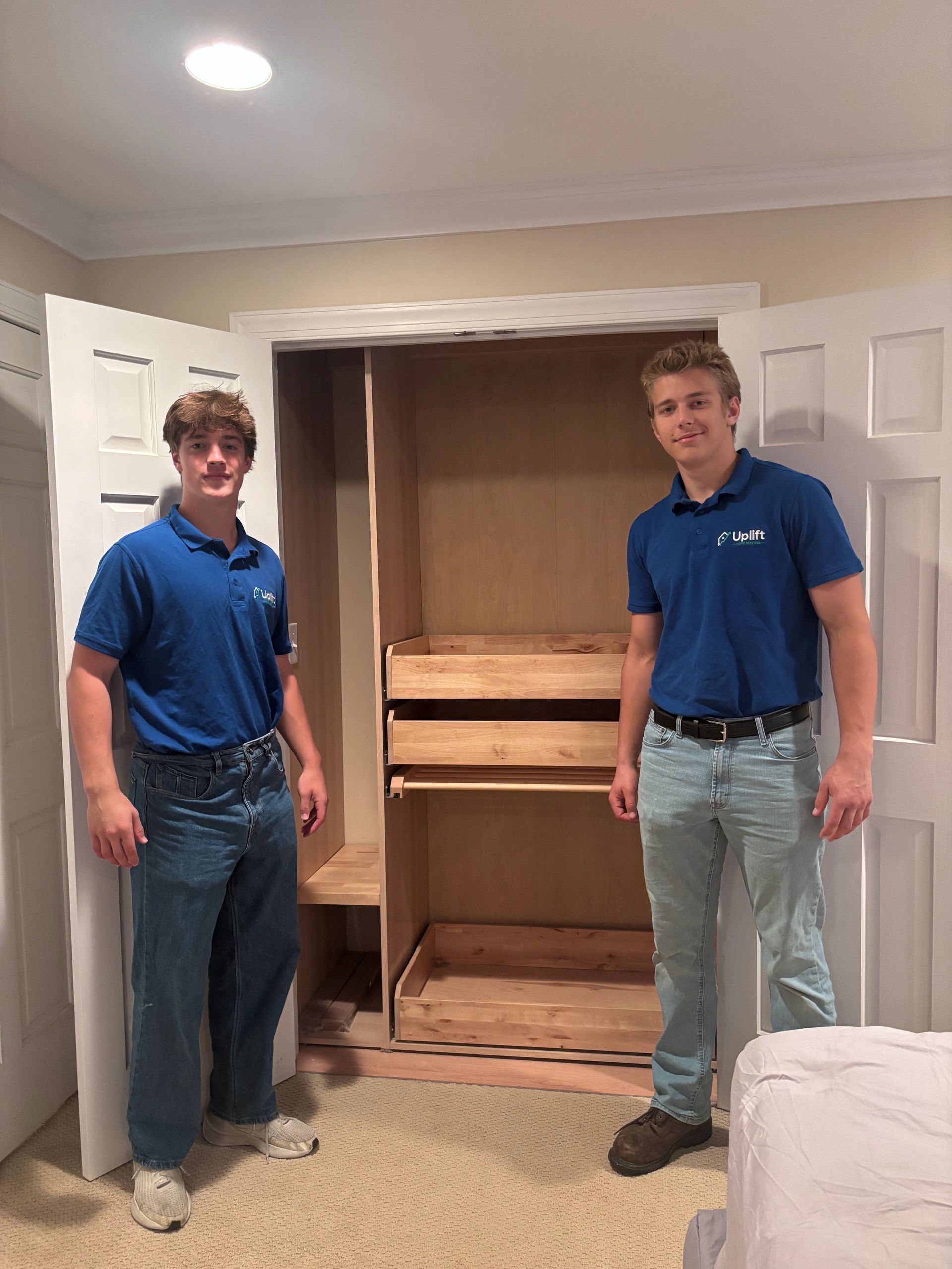 Two young men in blue shirts standing next to an open closet with wooden shelves.